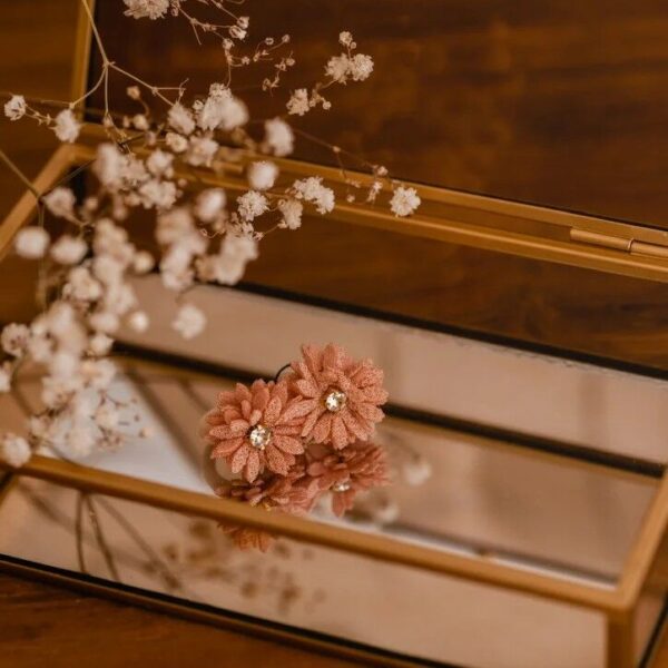 Twee oudroze elastiekjes met zachte bloemen en een klein sprankelend steentje in het midden, liggend in een gouden sieradendoos met spiegel. Droogbloemen op de achtergrond geven een warme, sfeervolle uitstraling.