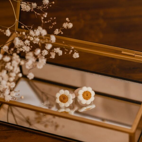 Een set van twee Blossom Daisy haarelastiekjes met geborduurde madeliefjes ligt op een spiegelend sieradendoosje. De zachte kleuren en verfijnde details geven de elastiekjes een speelse, maar stijlvolle uitstraling.