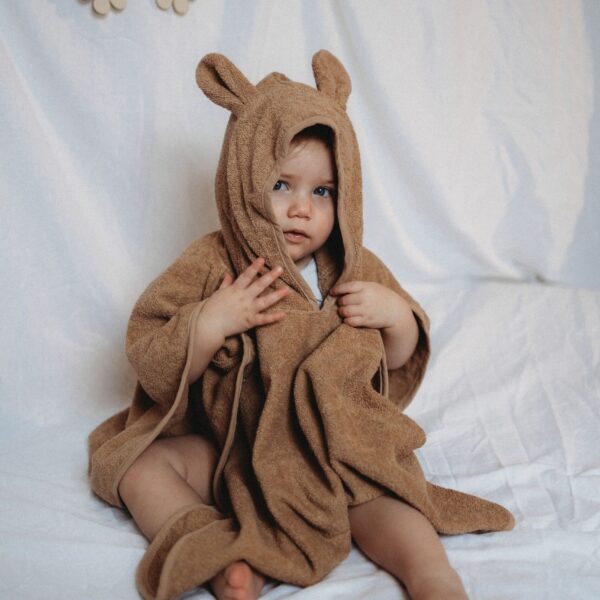 Baby in bruine badponcho met capuchon en naam Loïs geborduurd, zittend op wit beddengoed.