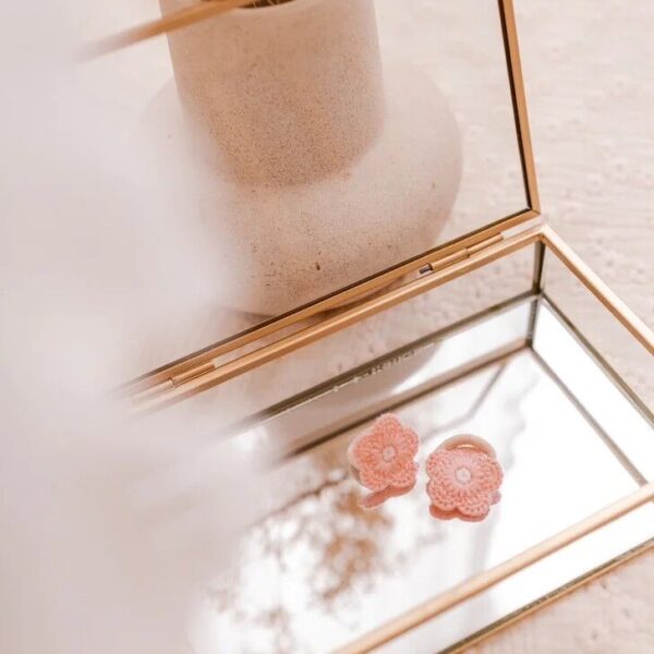Een setje van twee roze haarelastiekjes met geborduurde bloemen ligt op een spiegelend sieradendoosje met een gouden rand. De zachte tinten en natuurlijke styling geven de foto een rustige, elegante uitstraling.