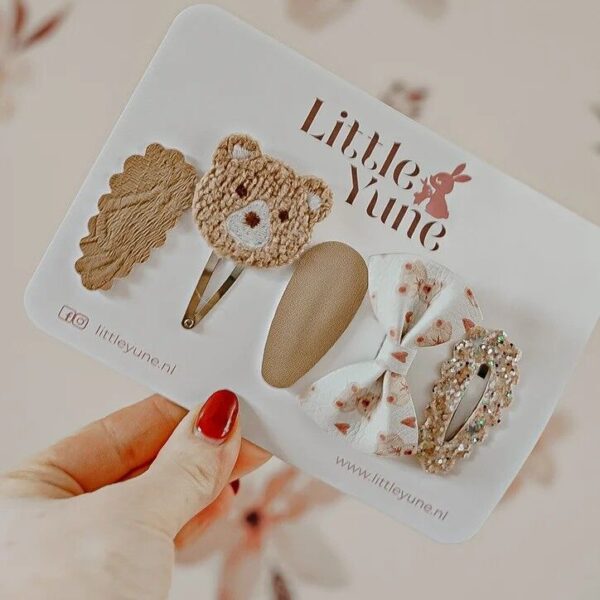 Een set van vijf haarspeldjes van Little Yune, genaamd 'Bear', met een mix van aardetinten, een geborduurd beertje, een strikje met berenprint en een glitterspeldje, gepresenteerd op een witte kaart tegen een zachte, neutrale achtergrond.