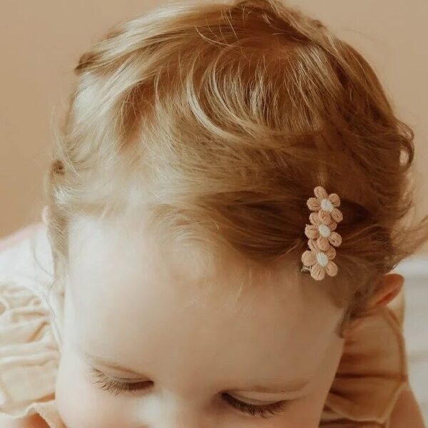 Een peuter met lichtblond haar draagt een setje daisy haarspeldjes, versierd met geborduurde bloemetjes in zachte tinten. De speldjes houden een klein plukje haar opzij en blijven stevig zitten dankzij de alligator grip. Ze draagt een luchtige outfit en lacht vrolijk.