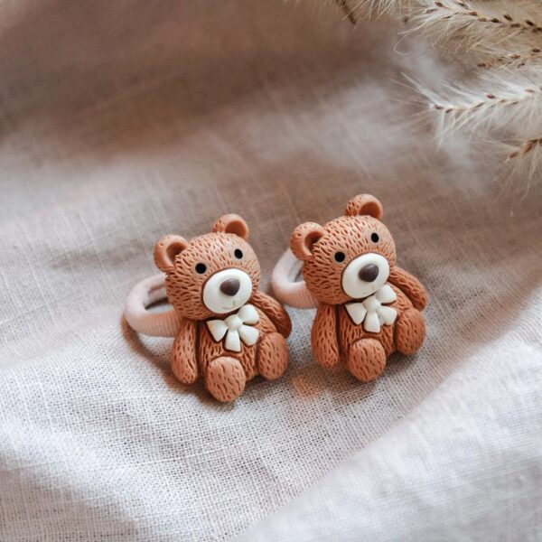 Twee schattige teddybeer-haarelastiekjes met strikje, gefotografeerd op een zachte linnen achtergrond.