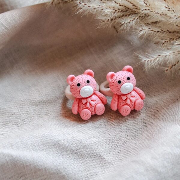 Twee roze teddybeer-haarelastiekjes met strikje, gefotografeerd op een zachte linnen achtergrond met pluimen.