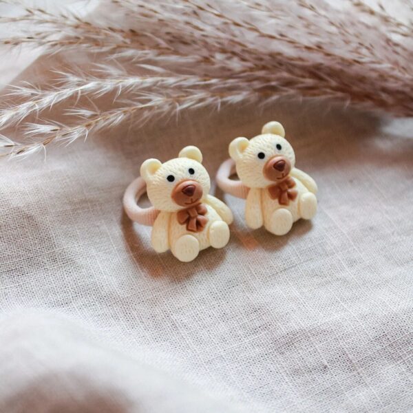 Twee crèmekleurige teddybeer-haarelastiekjes met bruin strikje, gefotografeerd op een zachte linnen achtergrond met pluimen.