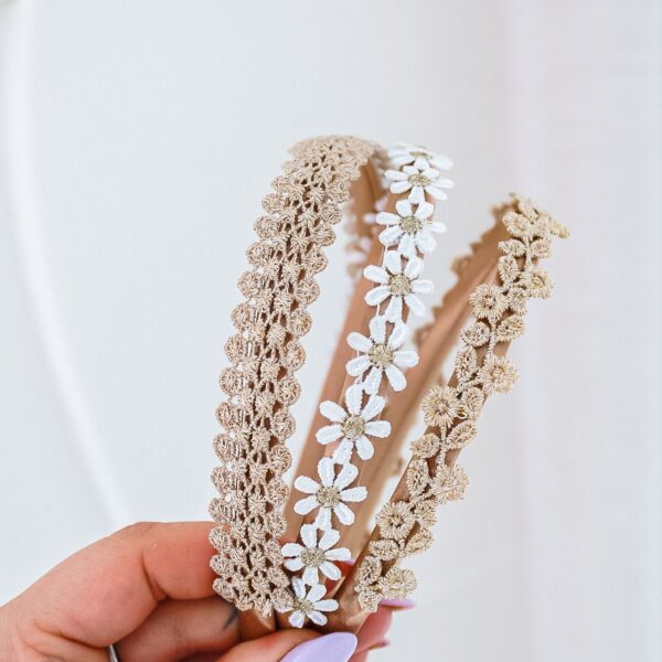 Close-up van drie geborduurde diademen in beige tinten met bloemenmotief, vastgehouden door een hand met lila nagellak.