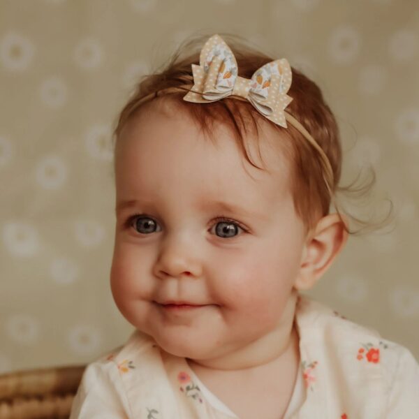 Baby meisje met lichtbruine haren draagt een beige paas haarbandje met stippen en konijntjesprint, zittend in een mandje met bloemenprint jurkje.