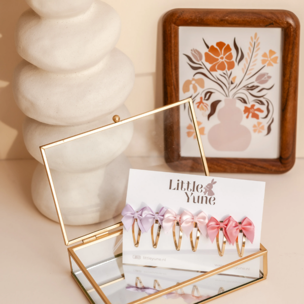 Set van zes satijnen strik haarspeldjes in zachte pasteltinten (paars, roze en lichtroze), bevestigd op 4 cm klikklakspeldjes, gepresenteerd in een sierlijke glazen doos met een bloemenvaas op de achtergrond. 🎀🌸