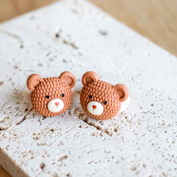 Twee karamelkleurige teddybeer-haar­elastiekjes op witte elastiek, liggend op een lichte steenachtige ondergrond.