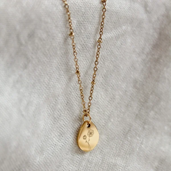 Close-up van een gouden geboortebloem ketting met gegraveerde bloem op een druppelvormige hanger, liggend op een zachte linnen achtergrond.