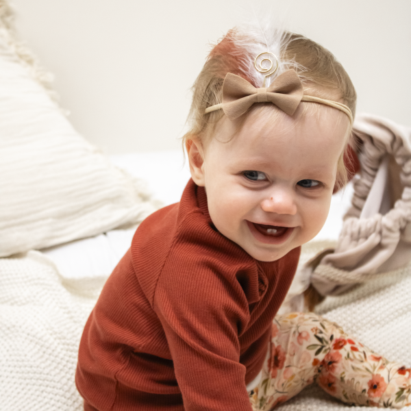 Baby met bruine strik en gouden veertje op een rekbaar pieten haarbandje, zittend in warme herfstoutfit op bed.