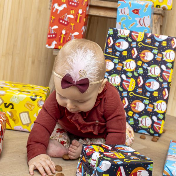 Baby met bordeauxrood haarbandje met strik en gouden veertje, zittend in feestelijke outfit met kruidnoten en sinterklaassfeer.