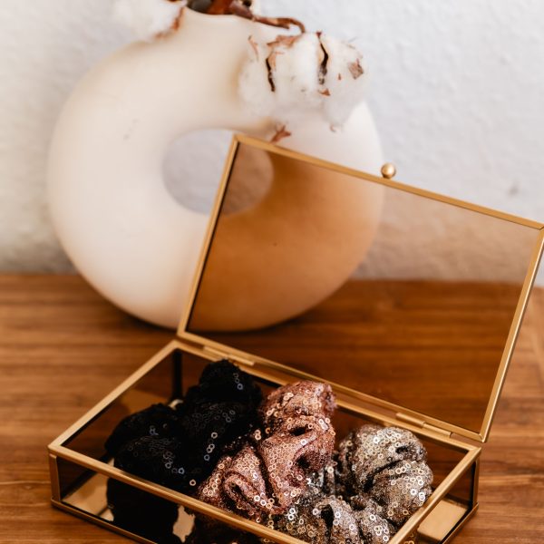 Scrunchie Luna van Little Yune – glanzende scrunchies met pailletten in zwart, roségoud, zilver en champagne, sfeervol gepresenteerd in gouden glazen doosje op houten tafel.