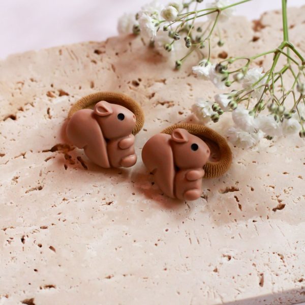 Twee schattige bruine elastiekjes met eekhoorntjes, liggend op beige steen met witte bloemetjes op de achtergrond.