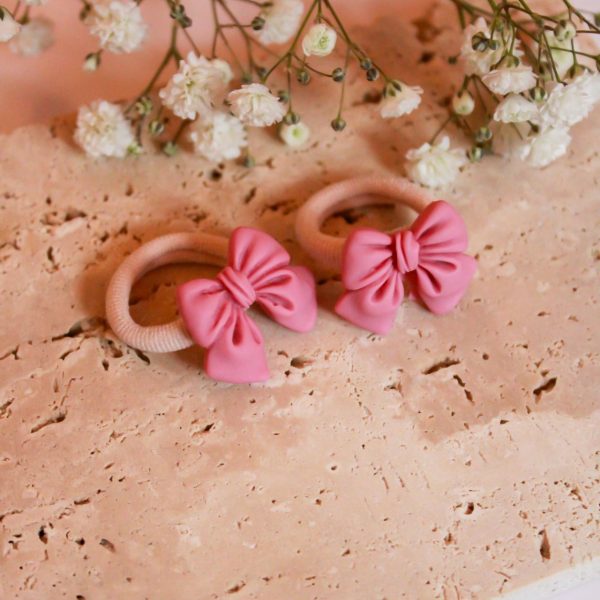 Twee roze strik-elastiekjes van Little Yune, liggend op een beige stenen ondergrond met witte bloemetjes erboven.