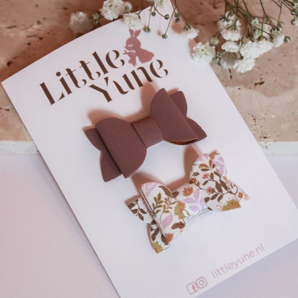 Twee antislip speldjes met strikjes: één in warme mauvetint en één met bloemenprint in zachte tinten, gepresenteerd op een Little Yune kaart met witte bloemetjes erboven.