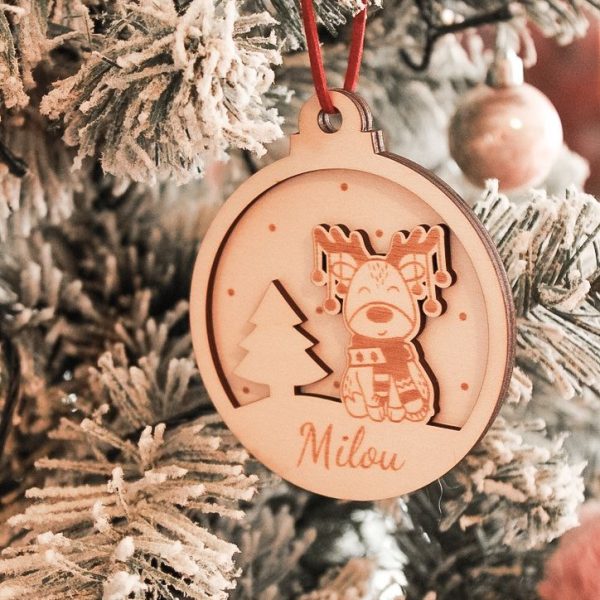 Houten kerstbal van Little Yune met gegraveerd rendiertje met sjaal en naam “Milou”, hangend in een besneeuwde kerstboom met rood suède koordje.