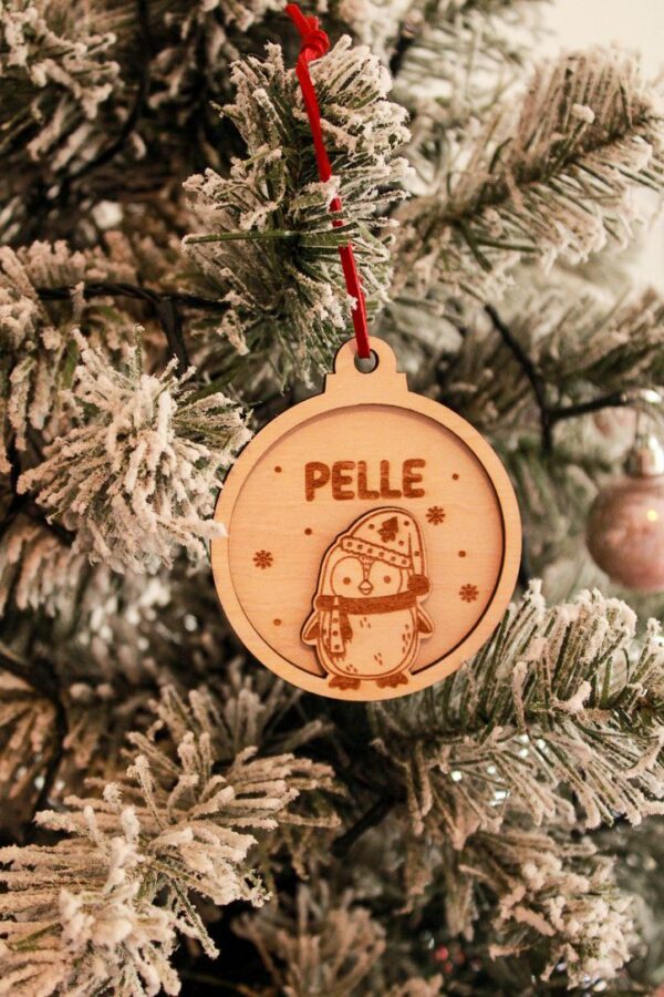 Houten kerstbal van Little Yune met gegraveerde pinguïn in muts en sjaal en naam ‘Pelle’, hangend in een besneeuwde kerstboom met rood suède koordje.