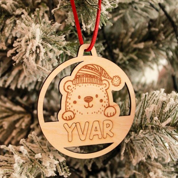Houten kerstbal van Little Yune met gegraveerd beertje met kerstmuts en naam “Yvar”, hangend in een besneeuwde kerstboom met rood suède koordje.