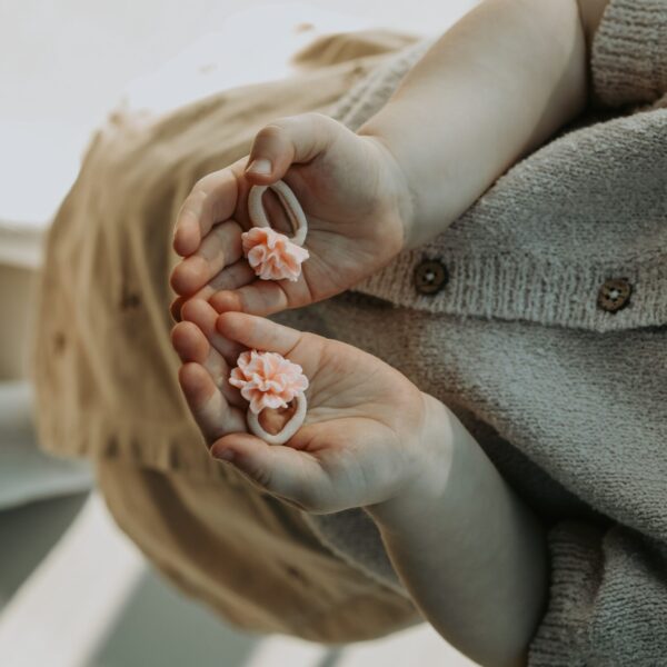 Set van twee pioenbloem elastiekjes in peachkleur op beige elastiek, vastgehouden in kinderhandjes.