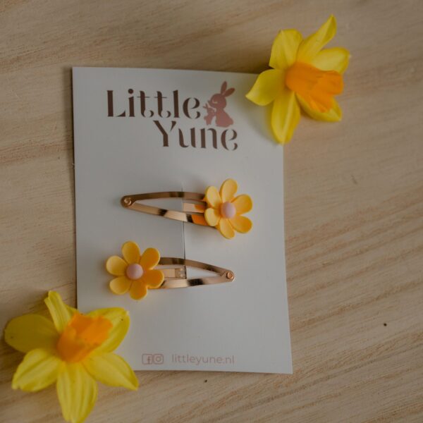 Twee gele madelief haarspeldjes op gouden 4 cm klikklakspeldjes van Little Yune, gefotografeerd tussen gele lentebloemen.