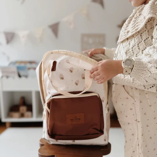 Crème rugzak van Mrs. Ertha met een vrolijk aardbeienprint, een warm bruin voorvak en bruine leren details — te zien op de rug van een jong meisje en geplaatst in een lichte kinderkamer naast een bijpassend lunchboxje.